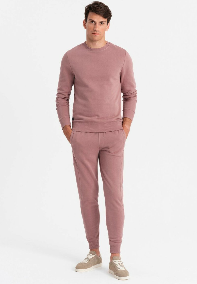 Helles rosa Sweatshirt und passende Jogginghose aus weichem Stoff, mit gerippten Bündchen und entspannter Passform. Graue Sneakers runden das Outfit ab.