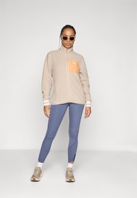 Fleecejakke i beige med en orange brystlomme, parret med blå leggings. Modellen bærer cremefarvede solbriller og beige sneakers.