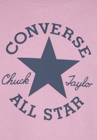 Felpa rosa con una stella blu e il testo "CONVERSE ALL STAR", con "Chuck Taylor" in un font corsivo. Tessuto in cotone morbido.