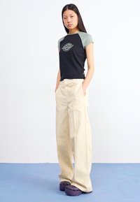 A young woman stands confidently, hands in pockets, wearing a black and gray shirt, beige trousers, and purple clogs against a plain background.