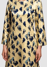 Vestido de manga larga en color beige con un estampado de aves estilizadas en azul, gris y negro, llevado por una persona con las manos relajadas a los lados.