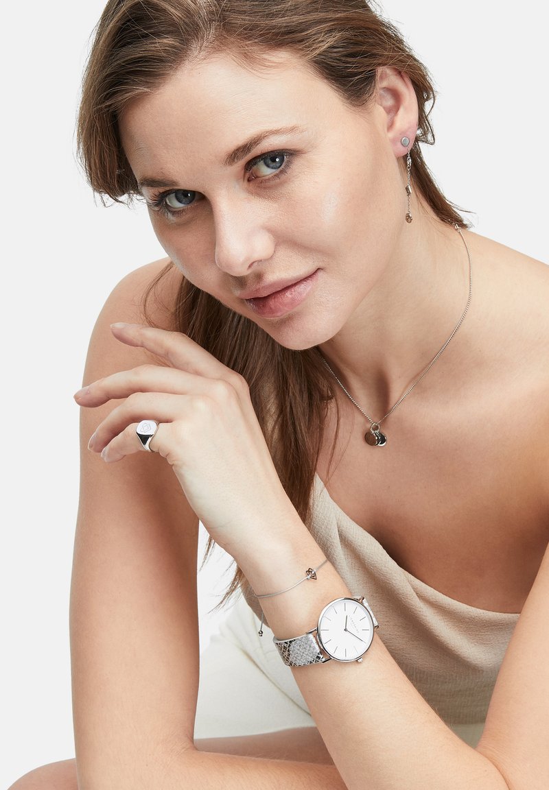 Reloj plateado con una correa gris texturizada, collares de plata en capas, pendientes de botón y un anillo geométrico. Ropa en colores neutros.