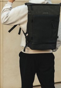 Musta rullatutti reppu, jossa on säädettävät olkahihnat, teksturoitu kangas, etutasku vetoketjulla ja logoyksityiskohtia yläosassa.