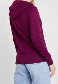 Personne portant un sweat à capuche prune à manches longues et un jean bleu clair, tourné de dos avec un bras détendu le long du corps.
