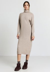 Robe tricotée beige avec un col roulé haut, des manches longues et une silhouette ajustée. Associée à des bottes à chevilles marron foncé et des chaussettes texturées.