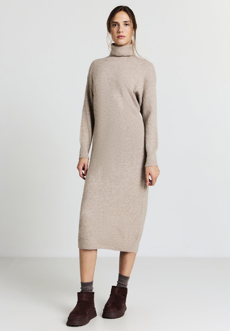 Robe tricotée beige avec un col roulé haut, des manches longues et une silhouette ajustée. Associée à des bottes à chevilles marron foncé et des chaussettes texturées.