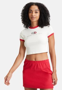 Camiseta corta blanca con ribete rojo, que presenta un logo negro, combinada con una falda mini de cintura elástica roja. Tela suave, diseño casual.