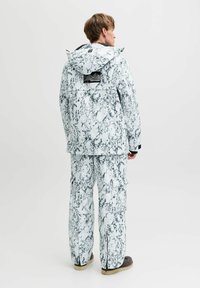 Personne portant une veste à capuche marbrée blanche et grise avec un pantalon cargo assorti, se tenant de dos sur un fond uni.