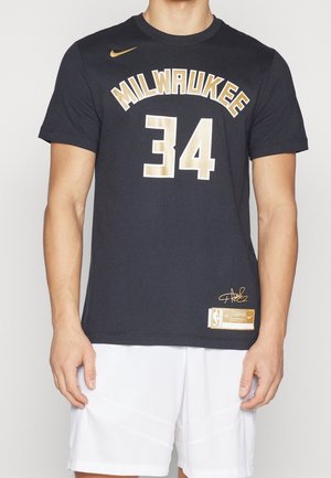 Mężczyzna w czarnej koszulce koszykarskiej Milwaukee z numerem 34 w złotym kolorze i białych szortach, stojący na jednolitym tle.
