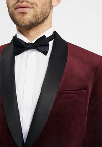 Burgundowy aksamitny marynarka z czarnym satynowym szalowym klapem i czarną muszką, noszona na białej koszuli z mankietami z kieszonką po lewej stronie.