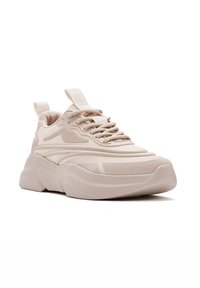 Beige Sneaker mit einer strukturierten Oberfläche, ausgestattet mit Schnürverschluss und einer Zuglasche. Dicke Sohle mit dezenten Rillen und runder Form.
