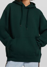 Felpa verde scuro con tasca frontale, cappuccio regolabile con lacci e polsini a coste. Materiale morbido con vestibilità rilassata.