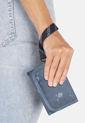 Hand hält ein blaues Stoffportemonnaie mit Blumenmuster und schwarzem Handgelenksriemen neben der Tasche einer hellblauen Jeans.