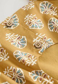 Blouse en coton jaune moutarde avec un motif floral blanc, bleu et marron. Dispose de deux boutons marron et d'un détail de patte de boutonnage cousue.