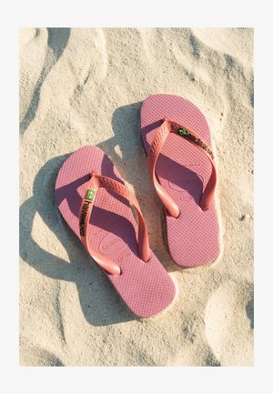 Enfant portant un short rose et des tongs, debout sur une plage de sable avec des parasols bleus, rouges et jaunes au bord de la mer.