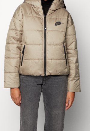 Kurtka puchowa w jasnym beżu, z kapturem, ciemnym zamkiem błyskawicznym i kieszeniami bocznymi. Posiada pikowane panele oraz czarne logo Nike na lewej piersi.