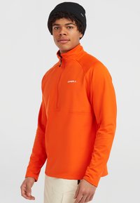 Oranje, rits fleece pullover met een hoge kraag, lichtgewicht materiaal en zwart merkembleem. Draagt een zwarte gebreide muts.