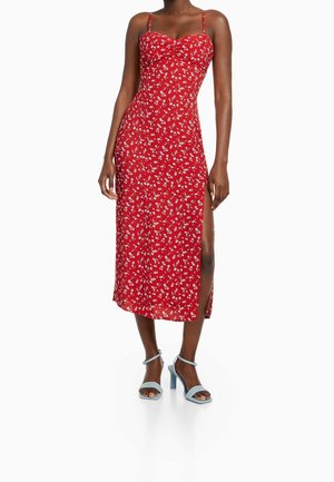 Czerwona midi sukienka w kwiaty z cienkimi ramiączkami, rozcięciem po boku i dopasowanym gorsetem. Posiada gładką teksturę i białe wzory kwiatowe. Zestawiona z niebieskimi szpilkami.