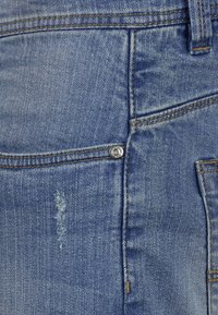 Jeans de mezclilla azul claro con una textura desgastada. Presenta un botón de metal y abrasiones sutiles en la tela. La costura es visible a lo largo de las costuras.