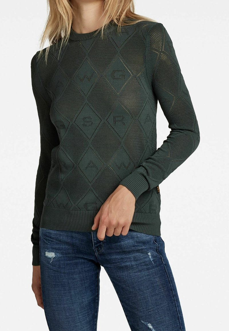 Suéter de punto verde oscuro con patrones de diamantes y letras texturizadas, diseño ajustado, y puños y dobladillo acanalados, combinado con jeans azules.