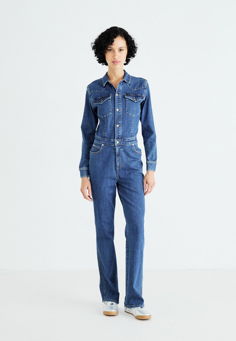 Combinaison en denim bleu moyen avec une coupe ajustée, des manches longues, deux poches poitrine et une fermeture à boutons à l'avant. Accents argentés à la taille.