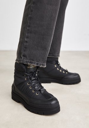 Bottes montantes noires avec des semelles épaisses et des œillets dorés, portées avec un jean gris foncé sur un sol clair.