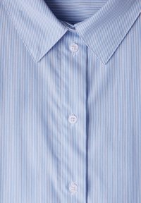 Camicia azzurra chiara con colletto classico, caratterizzata da strisce bianche verticali e cinque bottoni sul davanti. Tessuto liscio e lavorato.