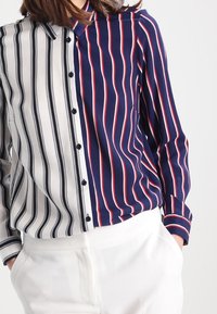 Koszula zapinana na guziki podzielona pionowo; lewa strona w czarno-białe paski, prawa strona w granatowe paski z czerwonymi i białymi. Długie rękawy i kołnierzyk.