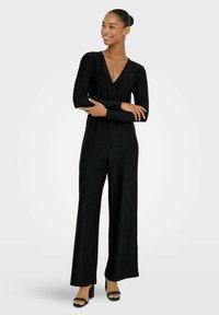 Mono negro de manga larga con escote en V, silueta holgada y un brillo sutil. Combinado con sandalias negras de tacón cuadrado.