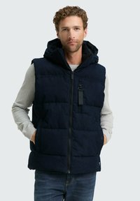 Vest puf cu glugă de culoare bleumarin, design matlasat, cu două buzunare laterale și închidere cu fermoar în față. Purtat peste o cămașă cu mânecă lungă de culoare gri deschis.