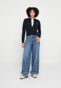 Navy knit cardigan with buttons, paired with wide-leg light blue denim jeans. Footwear features white ankle boots. Model stands against a plain background.