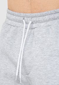 Pantalones de chándal grises de algodón con una cintura acanalada, que cuentan con cordones blancos y bolsillos laterales. El material tiene un acabado suave y texturizado.