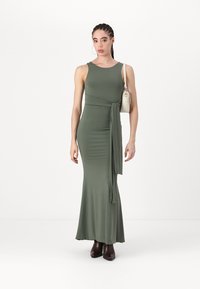 Robe maxi sans manches vert olive avec une silhouette ajustée, un détail enveloppant et une jupe fluide. Associée à des bottines brunes et un petit sac.