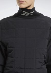 Top negro acolchado con cuello alto, con una superficie texturada y detalle del logotipo plateado en el cuello. El material parece ligero y elegante.