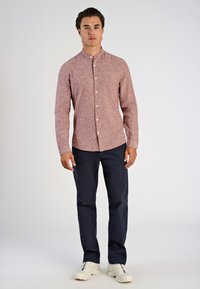 Camicia rossa a maniche lunghe con texture e colletto, abbinata a pantaloni blu navy e sneakers bianche. Presenta una tasca sul petto e bottoni bianchi.