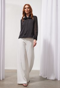 Black blouse with sheer polka dot sleeves and a bow tie detail, paired with loose white wide-leg trousers and black sandals.