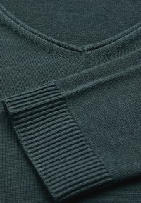 Ciemnozielony sweter dzianinowy z dekoltem w kształcie V i prążkowanymi mankietami, prezentujący gładką teksturę i równomierne przeszycie.