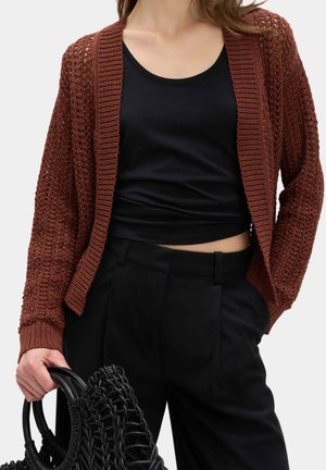Kobieta nosząca rdza-colored dzianinowy sweter na czarnym krótkim topie i czarnych wysokich spodniach, trzymająca czarną plecioną torebkę.