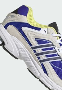 Kék és szürke sportcipő, hálós és velúrból készült, fehér díszítésekkel, textúrázott talppal és élénk sárga gallérral.