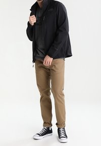 Schwarze wasserdichte Jacke über einem grauen T-Shirt, kombiniert mit khakifarbenen Hosen und schwarzen High-Top-Sneakern. Die Jacke verfügt über eine Reißverschlusstasche.