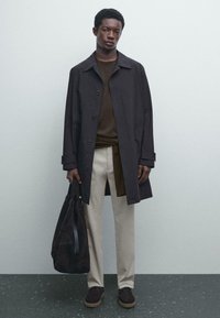 Mörkbrun trenchcoat med knäppning, bärs över en brun lagertröja. Beiga byxor, mörk väska och bruna skor fulländar looken.