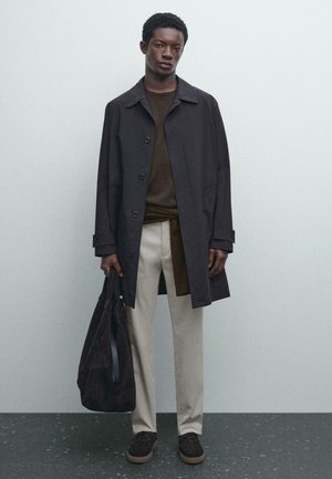 Man in een donkere jas, met een bruine shirt, lichtgekleurde broek en donkere schoenen, die een grote donkere tote bag vasthoudt tegen een effen achtergrond.