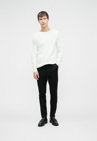 Bijela košulja s dugim rukavima, crne uske hlače i crne cipele, stojeći ispred jednostavne pozadine. Jednostavan, minimalistički dizajn.