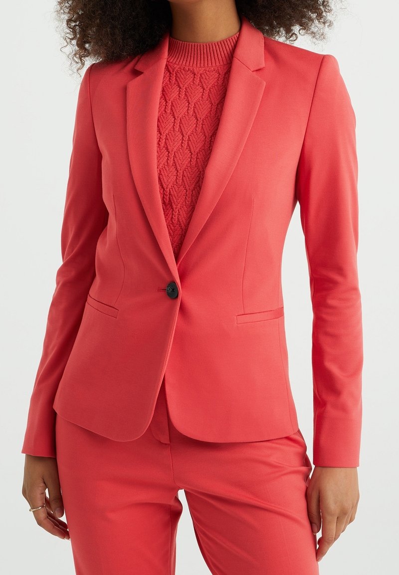 Femme portant un blazer ajusté corail avec un bouton noir sur un pull en maille corail texturé et un pantalon corail assorti.
