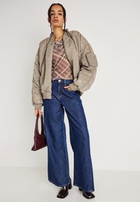 Casaco bomber verde oliva com punhos canelados, blusa ajustada de xadrez, jeans largos de denim azul e bolsa de pele roxa, combinados com botas escuras de salto.