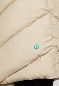 Beige Daunenjackenstoff mit einem gesteppten Design, das ein grünes Recycling-Logo als Akzent aufweist. Glatte Textur, sichtbare Nähte.
