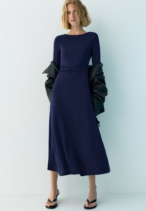 Vrouw in een lange marineblauwe jurk met een zwarte jas over haar armen gedrapeerd, staand tegen een effen lichte achtergrond.