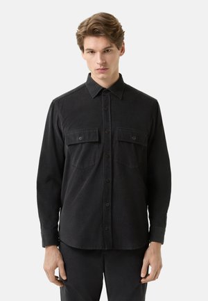 Ung mand iført en sort skjorte med knapper og to brystlommer, stående mod en ensartet lys baggrund.