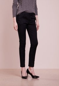 Schwarze maßgeschneiderte Hose mit schmaler Passform und umgeschlagenen Säumen, kombiniert mit einem grauen Strickpullover mit Zopfmuster und schwarzen High Heels vor neutralem Hintergrund.