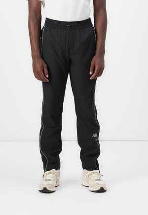 Black athletic pants with an elastic waistband, side stripes, and a New Balance logo. Worn with beige sneakers featuring dark soles.